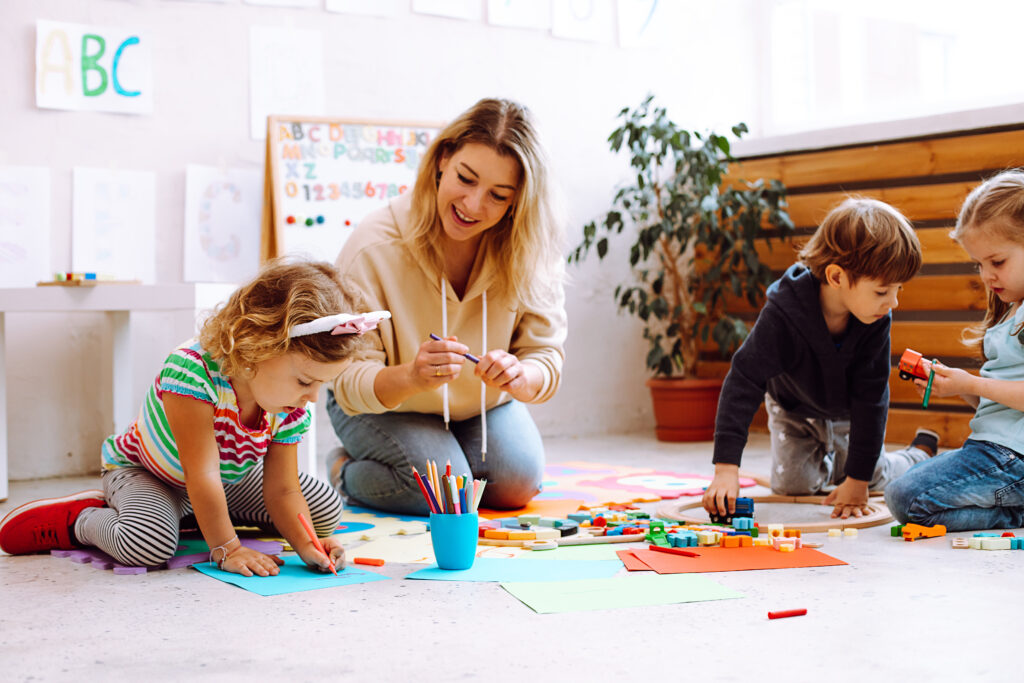 A smiling female teacher sits on the classroom floor with three young children engaged in creative activities—drawing, coloring, and playing with toys. Bright papers, crayons, and educational materials are scattered around, creating a playful and engaging preschool environment.